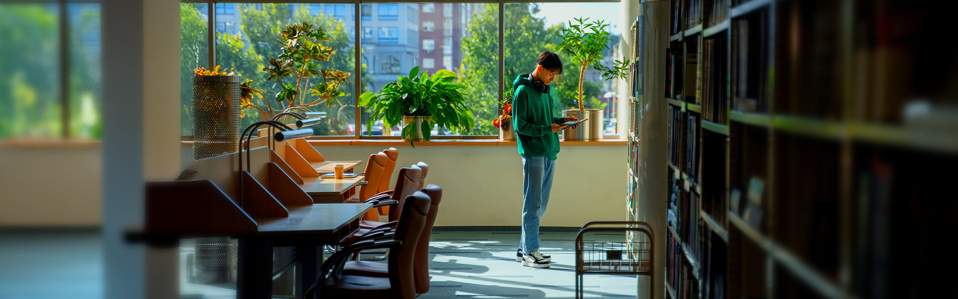 A guy standing in a library with large windows, plants, and bookshelves.
