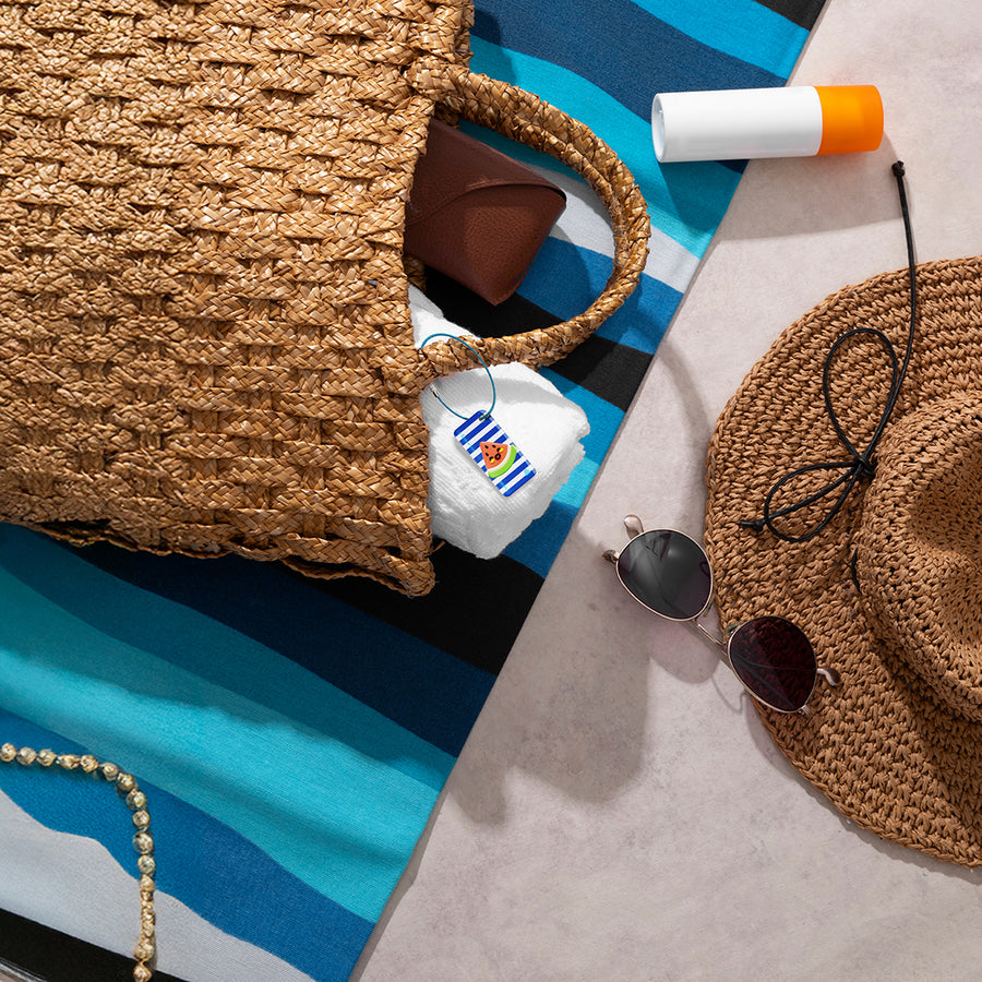 Straw bag with an nfc keychain attached to it, sunglasses, sunscreen and hat on a blue striped towel
